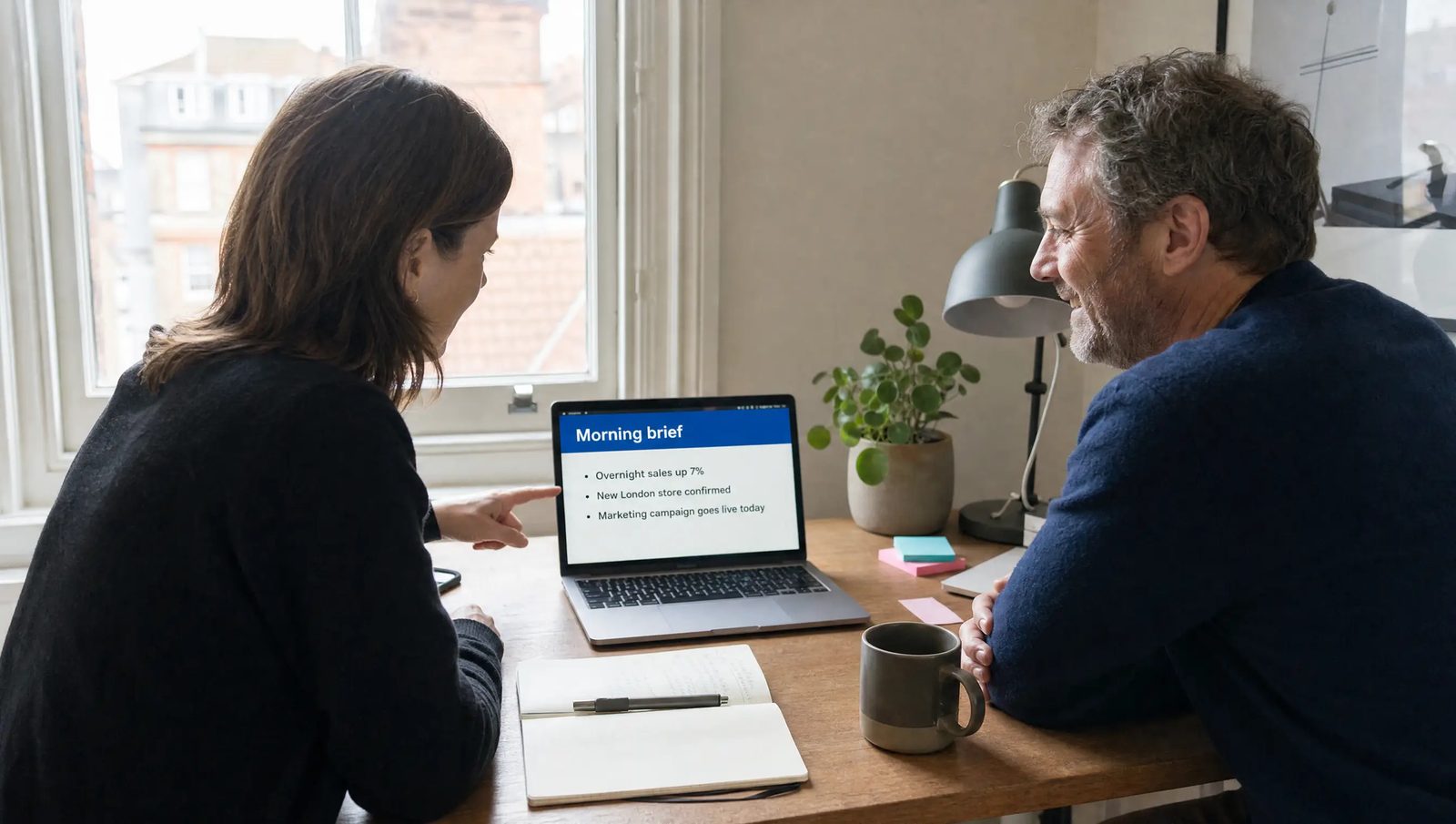 A woman and a man at a home-office desk in London, looking at a laptop showing a morning brief with 3 short lines.
