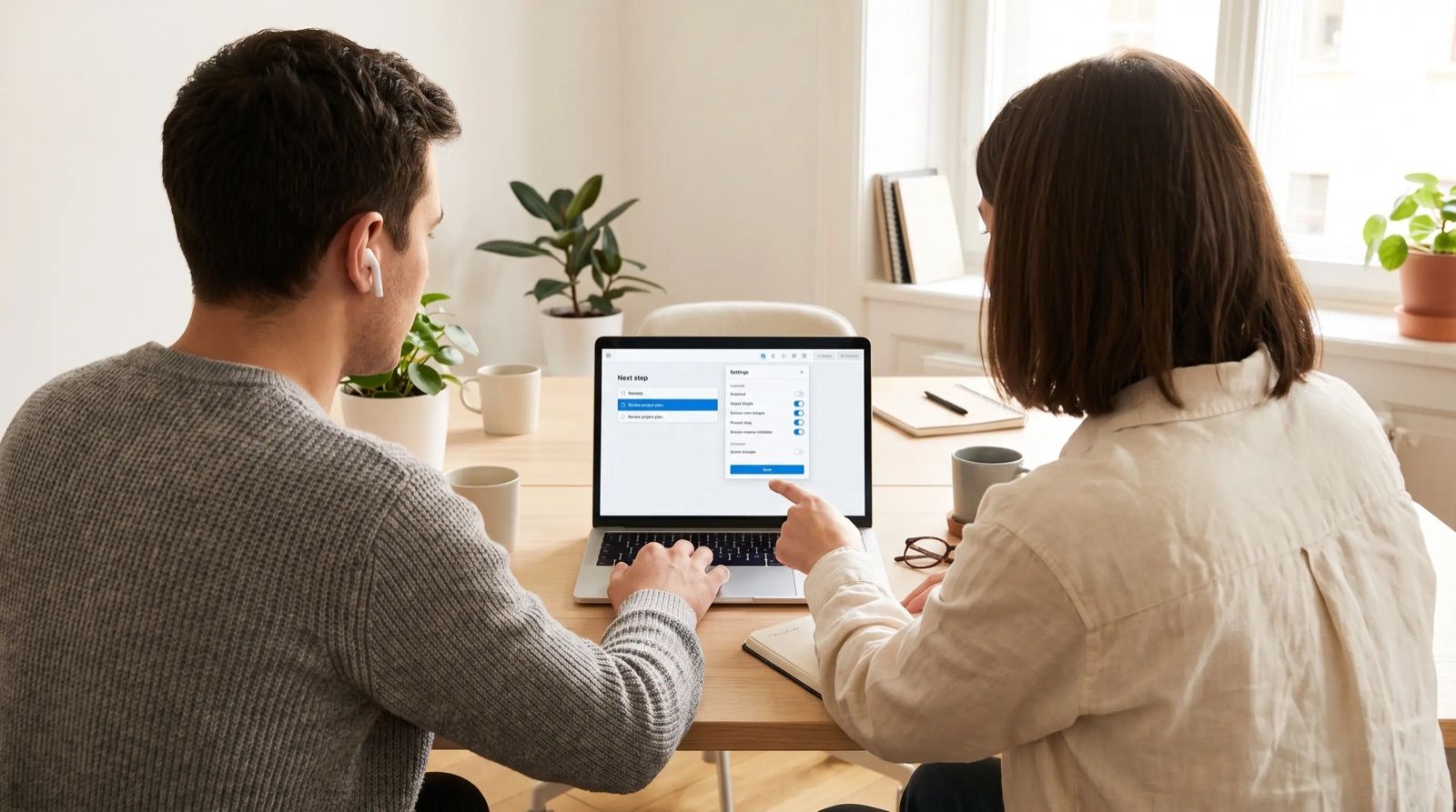 2 colleagues at a desk in a bright office, 1 wearing a wireless earbud, following a step shown on a laptop screen.